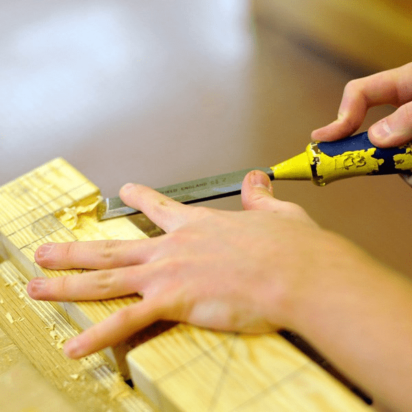 This photo depicts a person using a chisel on a piece of wood.
