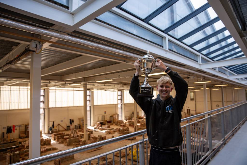 Image of Ellis Smith stood on the balcony overlooking the brick workshop holding the MPA Mortar trophy.
