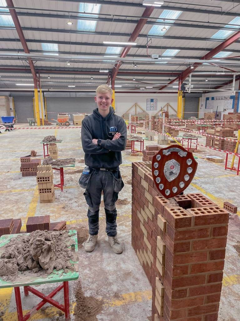 Image of Ellis Smith stood in a brick workshop next to his award winner's plaque