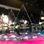 The photo features several glass trophies presented in rows on a table on the main stage of the award venue.