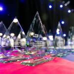 The photo features several glass trophies presented in rows on a table on the main stage of the award venue.