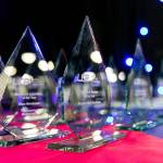 The photo features several glass trophies presented in rows on a table on the main stage of the award venue.