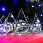 The photo features several glass trophies presented in rows on a table on the main stage of the award venue.