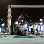 The photo features several glass trophies presented in rows on a table on the main stage of the award venue.