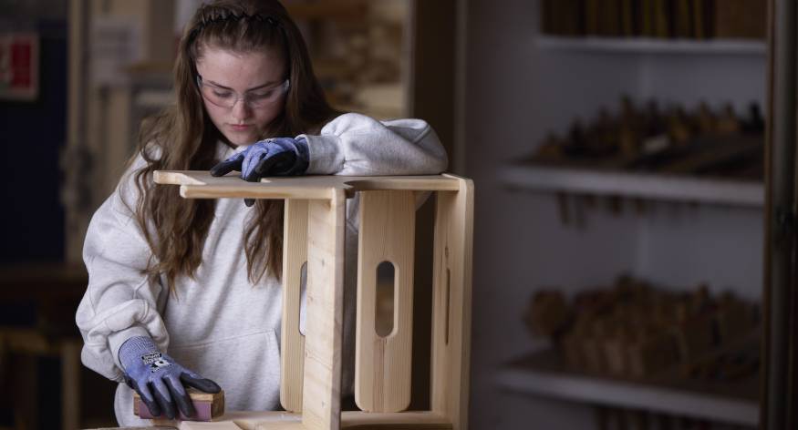 This image depicts a female LCB student sanding down a piece of wooden furniture.